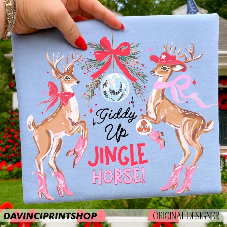 May include: Pale blue t-shirt with a festive design of two deer wearing cowboy boots and accessories. The text "Giddy Up Jingle Horse!" is in pink and white. A red bow and disco ball hang above the deer. The bottom of the shirt has the text "DAVINCIPRINTSHOP" and "ORIGINAL DESIGNER".