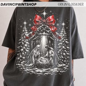 May include: A charcoal gray t-shirt featuring a graphic print of a nativity scene with Mary, Joseph, and the infant Jesus in a manger. The design includes two decorated Christmas trees, a large red bow at the top, and falling snowflakes.