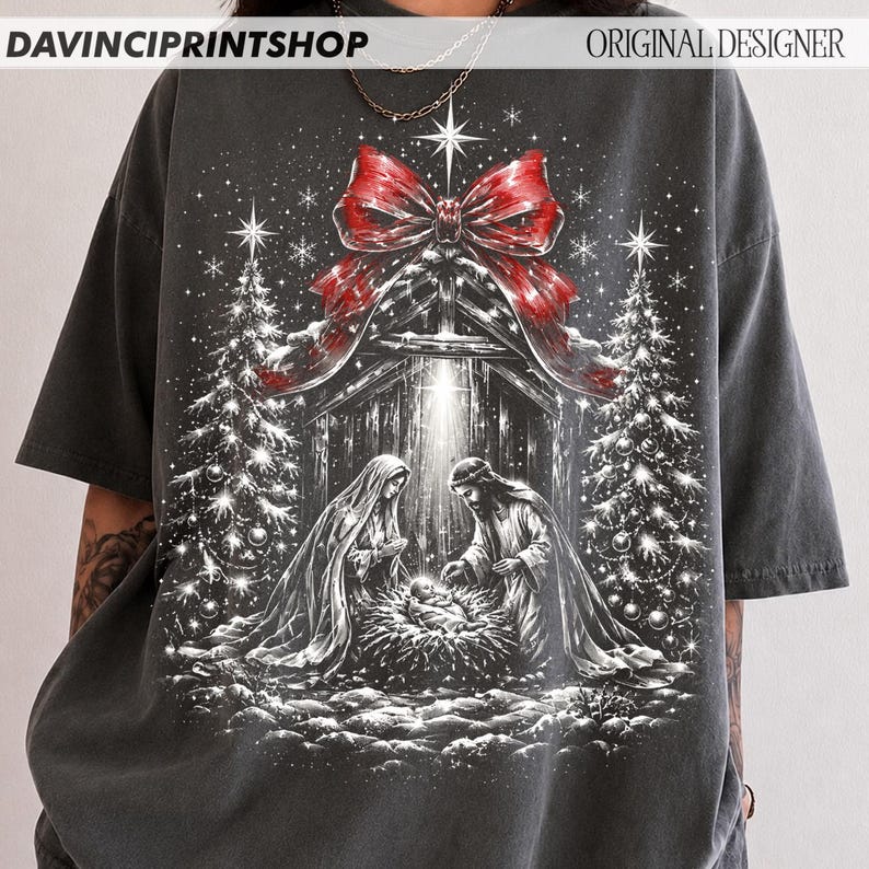 May include: A charcoal gray t-shirt featuring a graphic print of a nativity scene with Mary, Joseph, and the infant Jesus in a manger. The design includes two decorated Christmas trees, a large red bow at the top, and falling snowflakes.