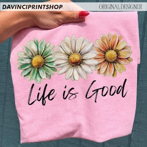 May include: A pink t-shirt with three daisy flower designs in green, white, and orange, each with a yellow centre. The words "Life is Good" are written in black script below the flowers. The shirt is held by a hand.