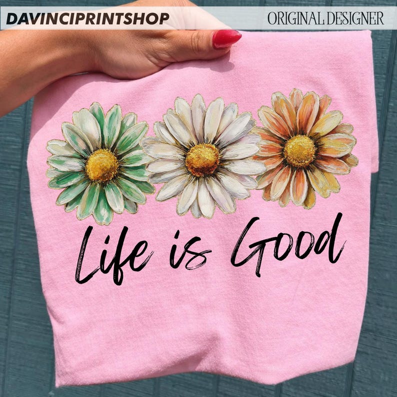 May include: A pink t-shirt with three daisy flower designs in green, white, and orange, each with a yellow centre. The words "Life is Good" are written in black script below the flowers. The shirt is held by a hand.
