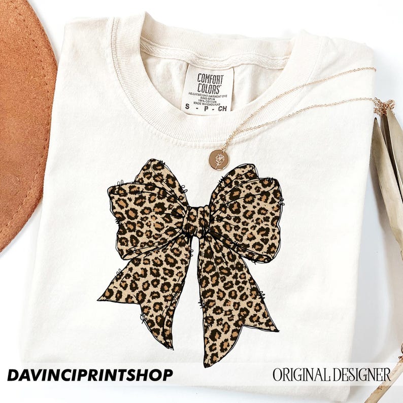 May include: A white t-shirt with a brown and black leopard print bow on the front. The shirt has a tag that says "Comfort Colors" and the text "DAVINCIPRINTSHOP" and "ORIGINAL DESIGNER" are at the bottom of the image.