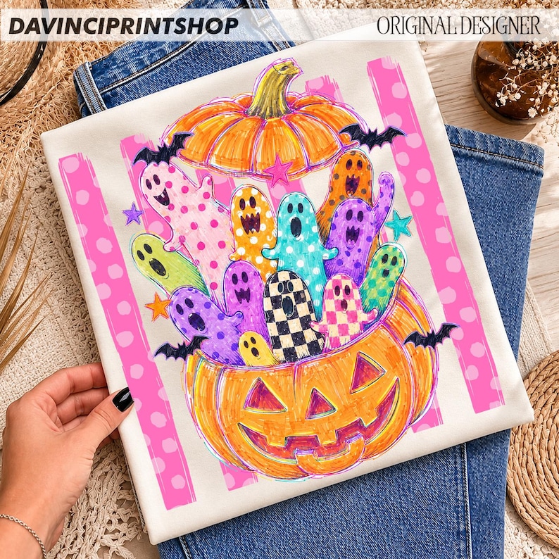 May include: A white t-shirt featuring a graphic print of a large orange jack-o'-lantern filled with colorful, patterned ghosts in shades of pink, yellow, blue, purple, and green, accented by small black bats and stars on a pink polka dot background.