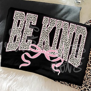Op de afbeelding: Zwarte stof met de woorden "BE KIND" in een luipaardprint lettertype met roze contouren. Een roze strik is gecentreerd onder de tekst. De achtergrond is een mix van zwart en luipaardprint.