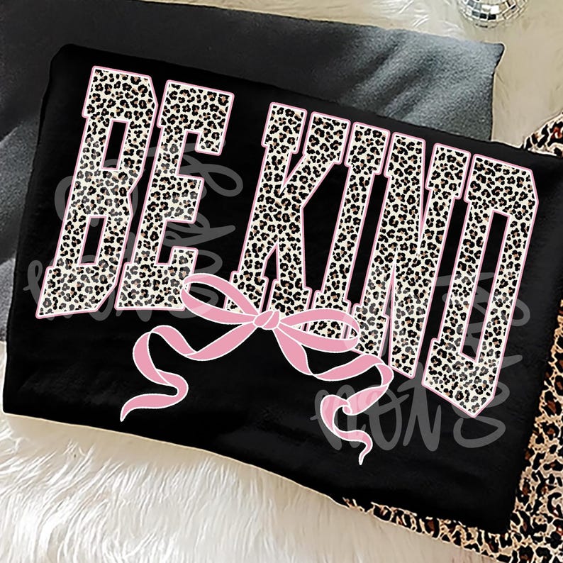 Op de afbeelding: Zwarte stof met de woorden "BE KIND" in een luipaardprint lettertype met roze contouren. Een roze strik is gecentreerd onder de tekst. De achtergrond is een mix van zwart en luipaardprint.