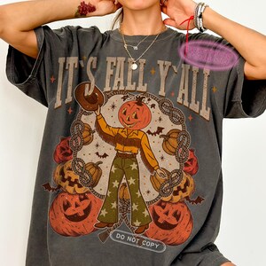 May include: Dark gray t-shirt with a pumpkin-headed cowboy graphic. The design includes the text "IT'S FALL Y'ALL" and a pumpkin-themed illustration with pumpkins, bats, and stars. The shirt also has the text "DO NOT COPY".