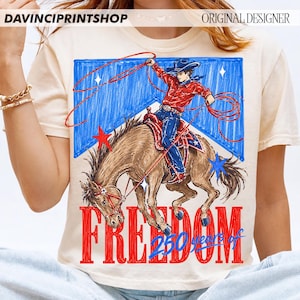 May include: Cream-colored t-shirt featuring a cowboy riding a horse with a lasso, set against a blue and white backdrop with stars. The word "FREEDOM" is in red, with "250 years of" below it. The shirt has the text "DAVINCIPRINTSHOP" and "ORIGINAL DESIGNER".