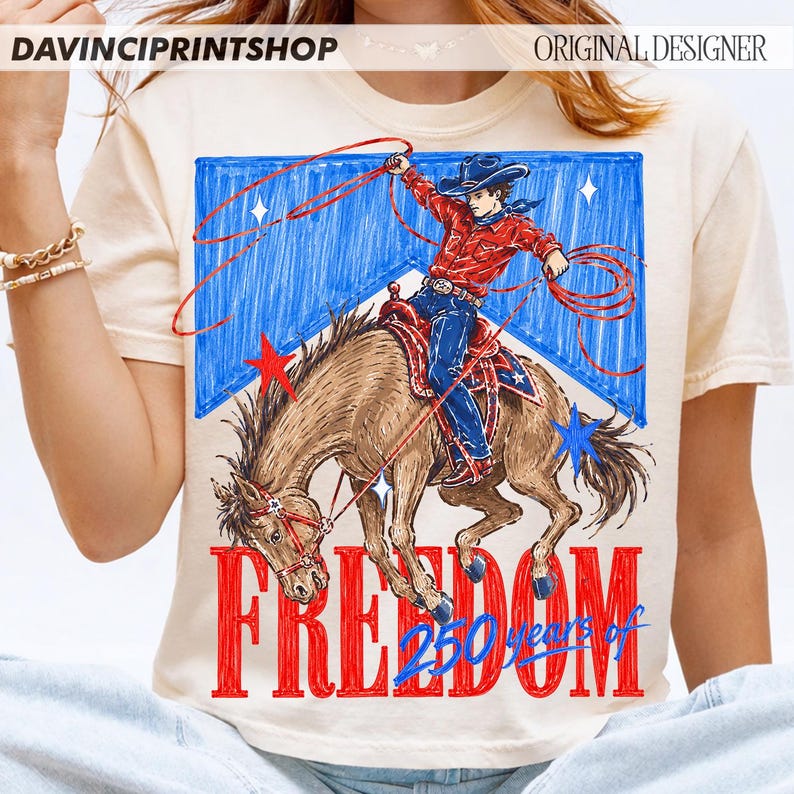 May include: Cream-colored t-shirt featuring a cowboy riding a horse with a lasso, set against a blue and white backdrop with stars. The word "FREEDOM" is in red, with "250 years of" below it. The shirt has the text "DAVINCIPRINTSHOP" and "ORIGINAL DESIGNER".