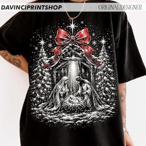 May include: A black t-shirt featuring a graphic print of a nativity scene with Mary, Joseph, and the infant Jesus. The design includes two decorated Christmas trees, a large red bow at the top, and a bright star, all set against a dark background.