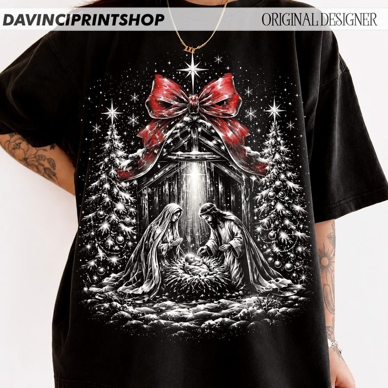 May include: A black t-shirt featuring a graphic print of a nativity scene with Mary, Joseph, and the infant Jesus. The design includes two decorated Christmas trees, a large red bow at the top, and a bright star, all set against a dark background.