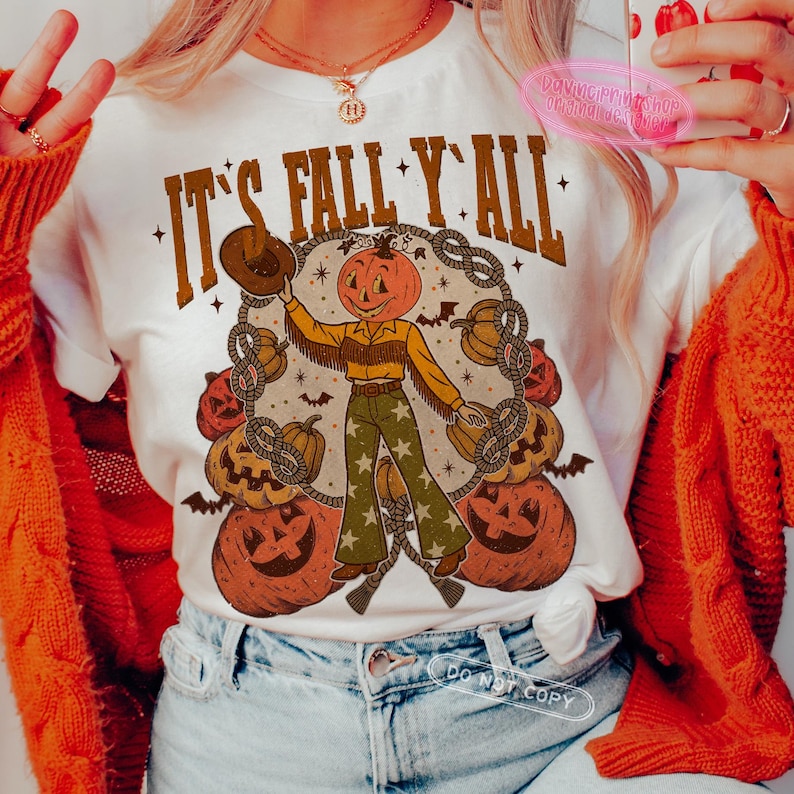 May include: White t-shirt with the text "IT'S FALL Y'ALL" in brown lettering. The shirt features a pumpkin-headed cowboy graphic surrounded by pumpkins and rope. The cowboy wears a yellow shirt, green pants with star patterns, and a brown hat.