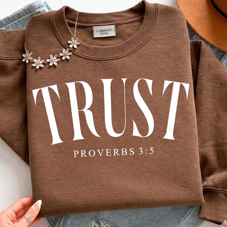 K&ouml;nnte beinhalten: Ein braunes Sweatshirt mit dem Wort "TRUST" in gro&szlig;en wei&szlig;en Buchstaben. Unter dem Wort "TRUST" steht der Text "PROVERBS 3:5" in kleineren wei&szlig;en Buchstaben. Eine Blumenkette ist &uuml;ber dem Sweatshirt drapiert.