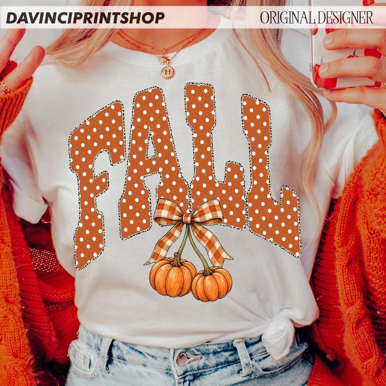 May include: A white t-shirt featuring the word FALL in large, orange letters with white polka dots. Below the text, two orange pumpkins are attached to a single stem with a brown and white checkered bow tied at the top of the pumpkin stems.