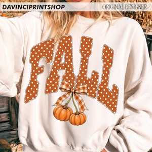 May include: A white long-sleeve sweatshirt featuring the word FALL in large orange letters with white polka dots and a leopard print outline. Below the text are two orange pumpkins connected by a brown and white checkered bow graphic.