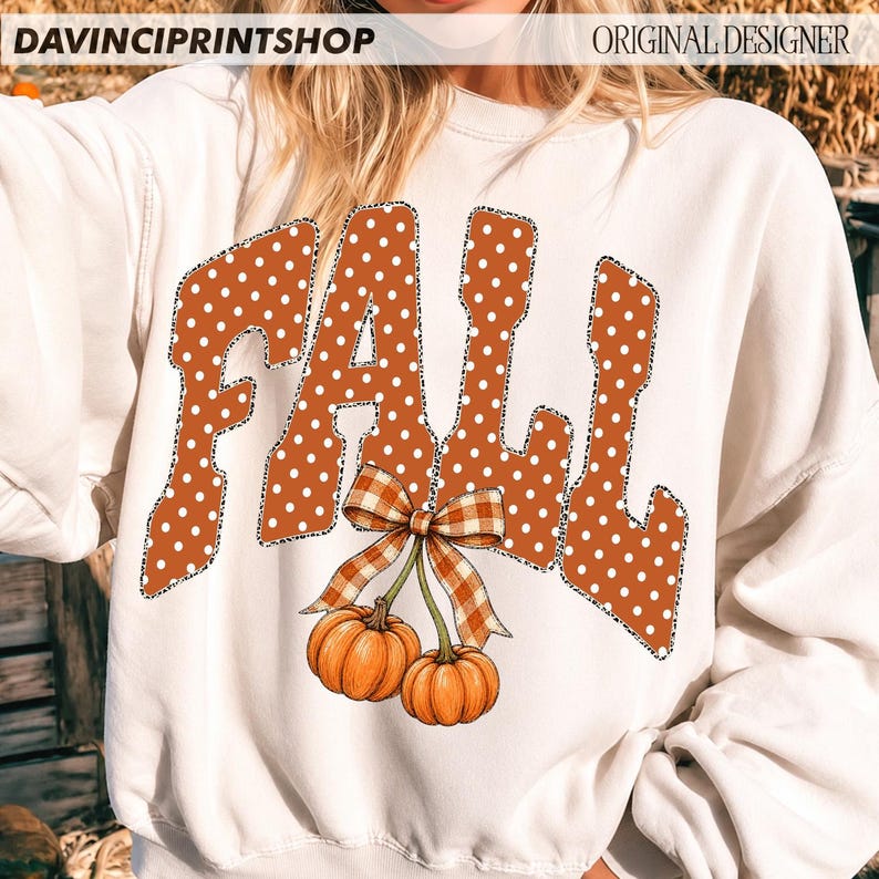 May include: A white long-sleeve sweatshirt featuring the word FALL in large orange letters with white polka dots and a leopard print outline. Below the text are two orange pumpkins connected by a brown and white checkered bow graphic.