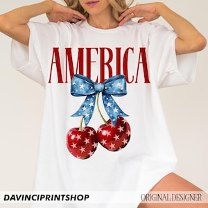 May include: White t-shirt with the word "AMERICA" printed in red letters. A blue bow with white stars is above the word, and two red cherries with white stars are below the bow.