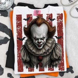 May include: A white t-shirt featuring a graphic of a clown with orange hair, a ruffled collar, and red makeup. The design includes vertical distressed red stripes and black bat silhouettes. The inner neck label reads Comfort Colors size L.
