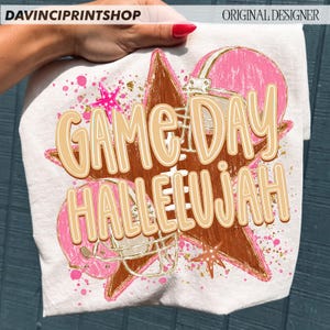 May include: A white t-shirt featuring a graphic print of a brown football, a pink football helmet, and pink stars. The design includes gold paint splatters and the text "GAME DAY HALLELUJAH" in large, light tan letters with a white outline.