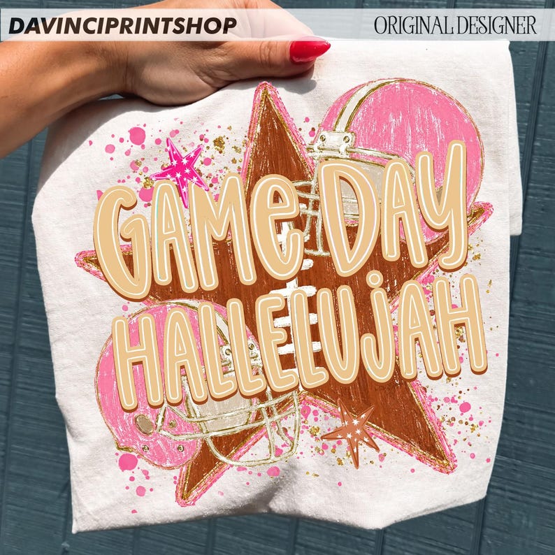 May include: A white t-shirt featuring a graphic print of a brown football, a pink football helmet, and pink stars. The design includes gold paint splatters and the text "GAME DAY HALLELUJAH" in large, light tan letters with a white outline.