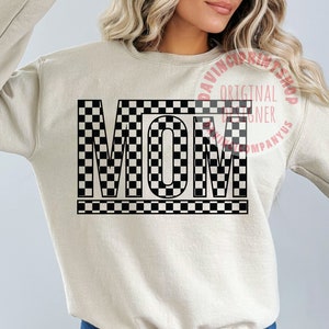 K&ouml;nnte beinhalten: Ein beiger Pullover mit dem Wort "MOM" in schwarz-wei&szlig;em Karomuster.