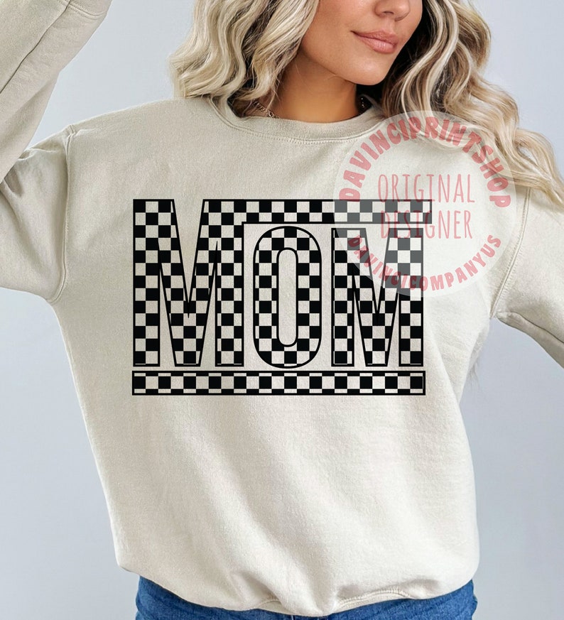 K&ouml;nnte beinhalten: Ein beiger Pullover mit dem Wort "MOM" in schwarz-wei&szlig;em Karomuster.