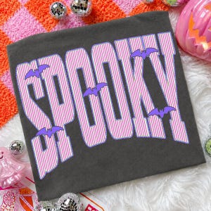 May include: A dark gray t-shirt featuring the word SPOOKY in large, bold letters. The letters have a pink and white vertical striped pattern with a purple outline. Several small, solid purple bat silhouettes are positioned around the text.
