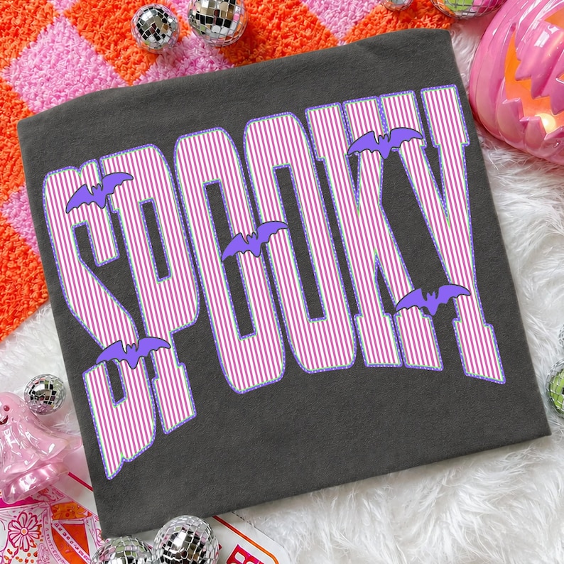 May include: A dark gray t-shirt featuring the word SPOOKY in large, bold letters. The letters have a pink and white vertical striped pattern with a purple outline. Several small, solid purple bat silhouettes are positioned around the text.