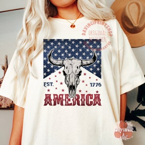 May include: A white t-shirt with a red, white, and blue graphic design. The graphic features a bull skull with horns, stars, and the text "EST. 1776 AMERICA".