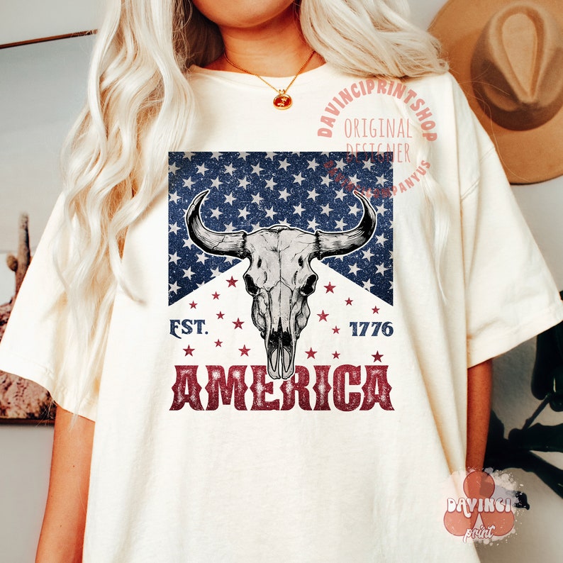 May include: A white t-shirt with a red, white, and blue graphic design. The graphic features a bull skull with horns, stars, and the text "EST. 1776 AMERICA".