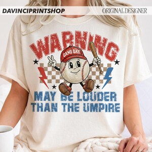 K&ouml;nnte beinhalten: Cremefarbenes T-Shirt mit Baseball-Motiv. Das Design zeigt einen Baseball-Charakter mit roter Kappe und Schl&auml;ger. Der Text "WARNING" und "MAY BE LOUDER THAN THE UMPIRE" ist in Rot und Blau gehalten.