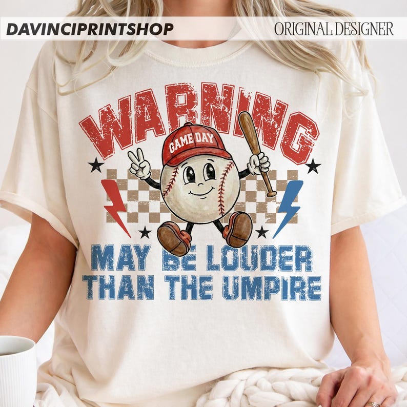 K&ouml;nnte beinhalten: Cremefarbenes T-Shirt mit Baseball-Motiv. Das Design zeigt einen Baseball-Charakter mit roter Kappe und Schl&auml;ger. Der Text "WARNING" und "MAY BE LOUDER THAN THE UMPIRE" ist in Rot und Blau gehalten.