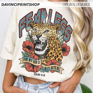 Puede incluir: Camiseta color crema con un gr&aacute;fico de leopardo y la palabra "FEARLESS" en letras turquesas y rojas. El dise&ntilde;o incluye amapolas rojas y el texto "FOR MY GOD IS ALWAYS WITH ME" e "ISAIAH 41:10".
