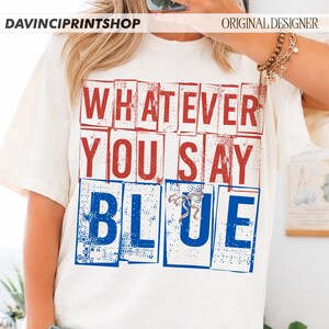 以下が含まれることがあります： クリーム色のTシャツ。赤と青のブロック体で「WHATEVER YOU SAY BLUE」と書かれています。デザインには小さなシルバーのリボンが含まれています。カジュアルな普段着です。