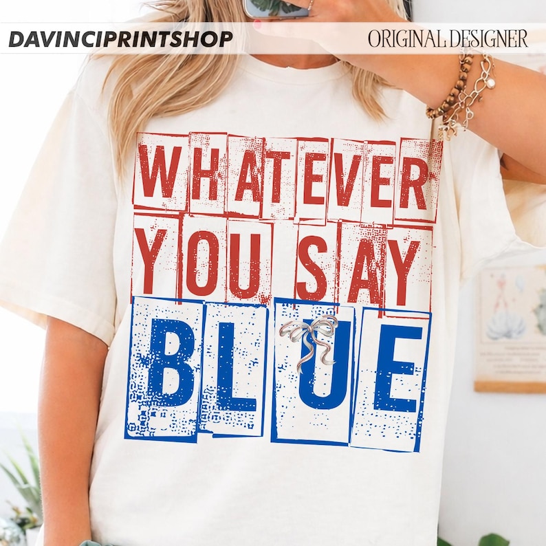 以下が含まれることがあります： クリーム色のTシャツ。赤と青のブロック体で「WHATEVER YOU SAY BLUE」と書かれています。デザインには小さなシルバーのリボンが含まれています。カジュアルな普段着です。