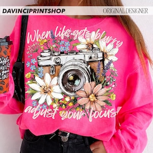 May include: A bright pink sweatshirt featuring a vintage camera design surrounded by flowers. The text "When life gets blurry, adjust your focus!" is written in white script. The sweatshirt has long sleeves and a crew neck.