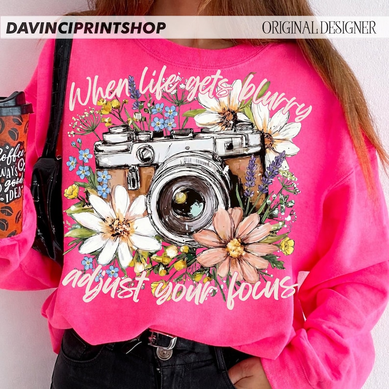May include: A bright pink sweatshirt featuring a vintage camera design surrounded by flowers. The text "When life gets blurry, adjust your focus!" is written in white script. The sweatshirt has long sleeves and a crew neck.