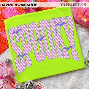 May include: A neon green t-shirt featuring the word SPOOKY printed in large, bold letters with a white and purple vertical striped pattern. Small purple bat silhouettes are positioned between and around the letters of the text.