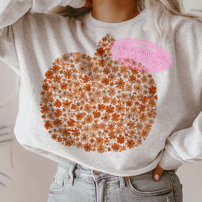 May include: A light gray sweatshirt featuring a pumpkin design made of orange and peach-colored flowers. The design is centered on the front of the sweatshirt. The text "Davinci print shop original designer" is on a pink oval.
