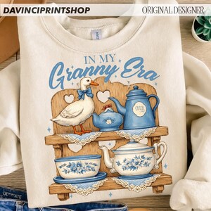 Puede incluir: Sudadera color crema con una ilustraci&oacute;n caprichosa. Un pato blanco con un lazo azul est&aacute; cerca de una estanter&iacute;a de madera con teteras azules y un cuenco. El texto "IN MY Granny Era" est&aacute; escrito en azul.