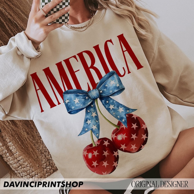 May include: A cream-colored sweatshirt with the word "AMERICA" printed in red letters. The sweatshirt features a blue and white bow with stars and two red cherries with white stars.