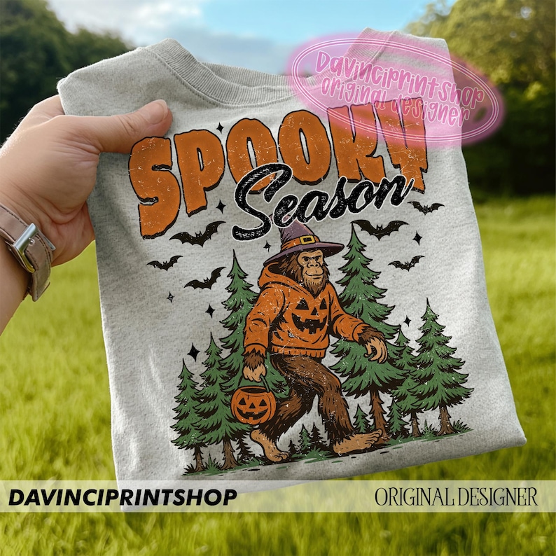 May include: Light gray sweatshirt with the words "SPOOKY Season" in orange and black. The graphic features a Bigfoot character wearing a pumpkin-themed hoodie and holding a jack-o'-lantern, with bats and pine trees. "DAVINCIPRINTSHOP" and "ORIGINAL DESIGNER" are printed at the bottom.
