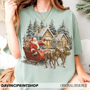 May include: A pale green t-shirt with a festive Christmas design. It shows Santa Claus in a red sleigh, pulled by reindeer, against a backdrop of snow-covered houses and fir trees. The text "DAVINCIPRINTSHOP" and "ORIGINAL DESIGNER" are at the bottom.