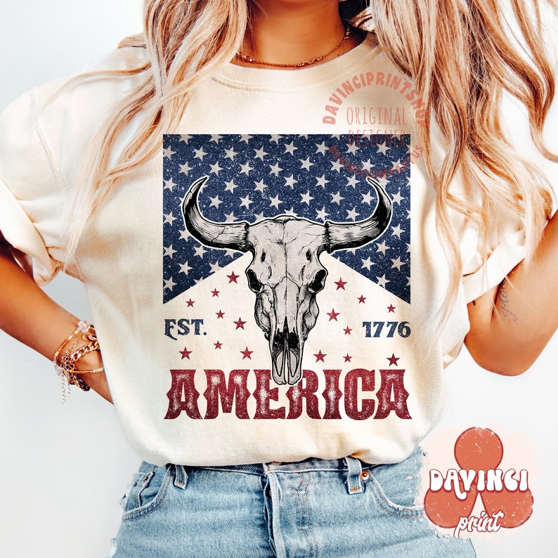 May include: A white t-shirt with a vintage-style graphic featuring a bull skull with a red, white, and blue American flag background. The text "EST. 1776 AMERICA" is printed below the skull.