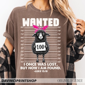Puede incluir: Una camiseta marr&oacute;n con una oveja negra que lleva un pa&ntilde;uelo rosa y sostiene un cartel que dice "100". La camiseta tiene un cartel blanco "WANTED" en la parte superior y el texto "I once was lost, but now I am found. -Luke 15:10" en la parte inferior.