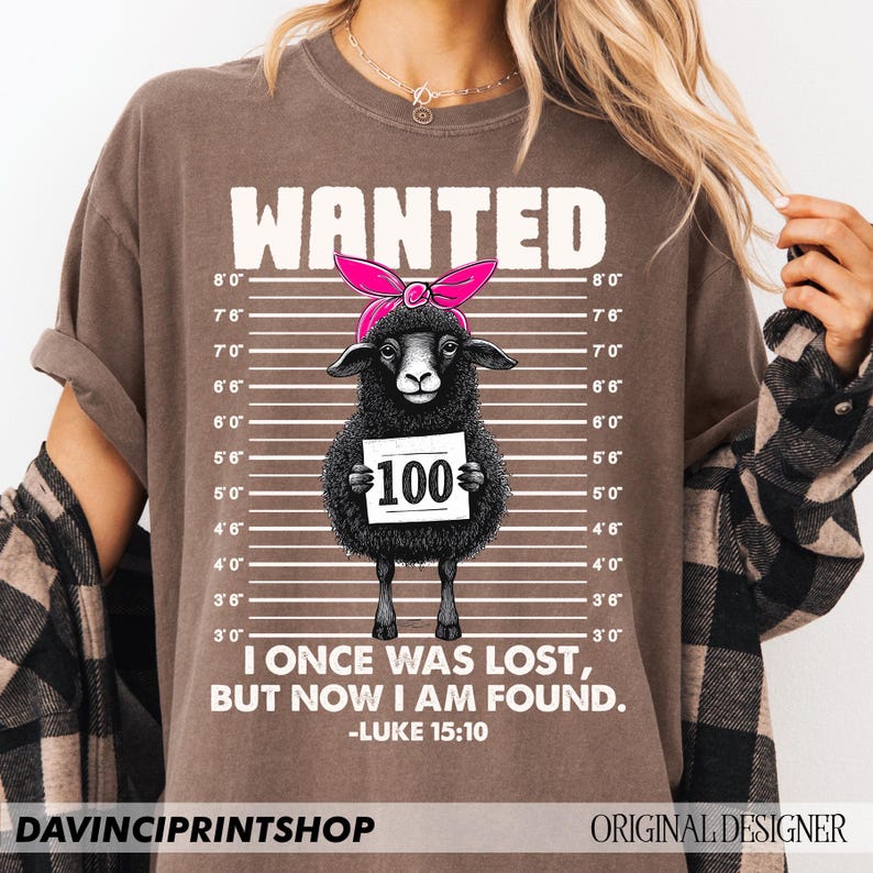 Puede incluir: Una camiseta marr&oacute;n con una oveja negra que lleva un pa&ntilde;uelo rosa y sostiene un cartel que dice "100". La camiseta tiene un cartel blanco "WANTED" en la parte superior y el texto "I once was lost, but now I am found. -Luke 15:10" en la parte inferior.