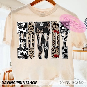 May include: Cream-colored t-shirt with the word "HOWDY" in a bold, patterned design. The letters feature cow print, leopard print, stars, and cherries. The shirt has short sleeves and a crew neck. The text "DAVINCIPRINTSHOP" is at the bottom.