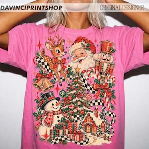 May include: A bright pink t-shirt featuring a festive Christmas graphic with a reindeer, Santa Claus, a nutcracker, a snowman, a decorated tree, wrapped gifts, bells, and a small house. The text DAVINCIPRINTSHOP and ORIGINAL DESIGNER appears at the top.