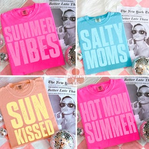 以下が含まれることがあります： 夏をテーマにしたテキストがプリントされた4枚のカラフルなTシャツ。ホットピンクのシャツには「Summer Vibes」、ライトブルーのシャツには「Salty Moms」、ピーチ色のシャツには「Sun Kissed」、ホットピンクのシャツには「Hot Mom Summer」と書かれています。各シャツはヴィンテージスタイルです。