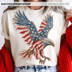 Pu&ograve; includere: T-shirt color crema con una grafica dettagliata di un'aquila calva americana con le ali spiegate. Il design dell'aquila incorpora elementi della bandiera americana, con toppe blu, rosse e bianche. I testi "DAVINCIPRINTSHOP" e "ORIGINAL DESIGNER" sono visibili.