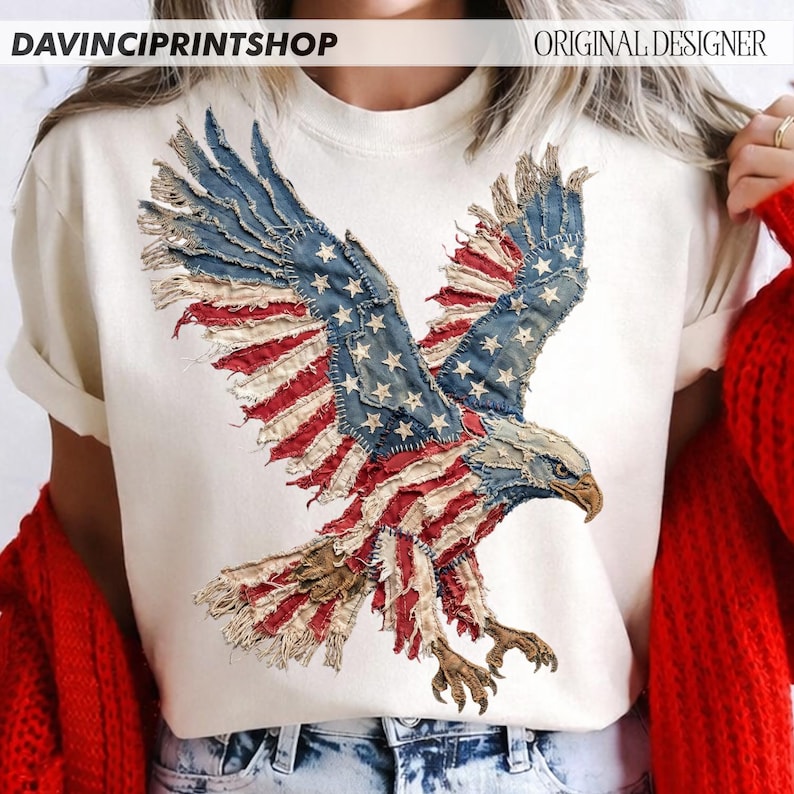 Pu&ograve; includere: T-shirt color crema con una grafica dettagliata di un'aquila calva americana con le ali spiegate. Il design dell'aquila incorpora elementi della bandiera americana, con toppe blu, rosse e bianche. I testi "DAVINCIPRINTSHOP" e "ORIGINAL DESIGNER" sono visibili.